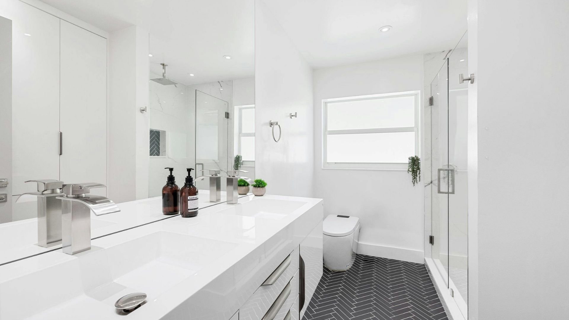 Modern white bathroom with double vanity, glass shower enclosure, and herringbone tile flooring.