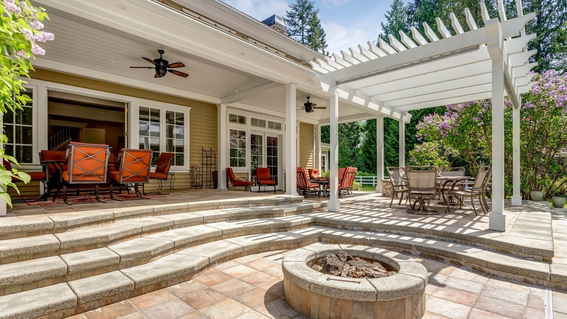 Luxury patio with white pergola, firepit, and orange seating under sunny clear sky.