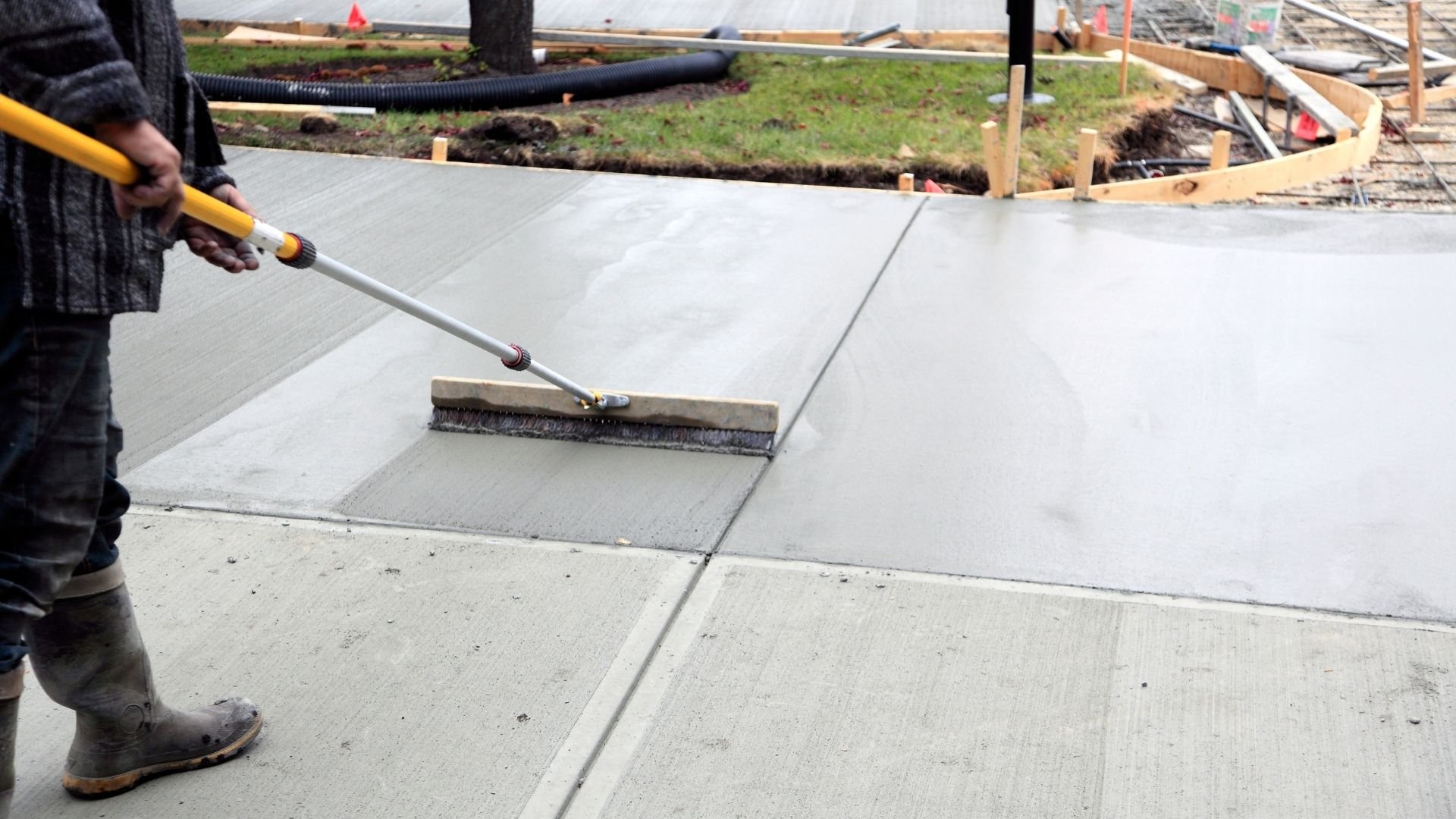 Worker using long-handled brush to smooth wet concrete slab during construction finishing work.